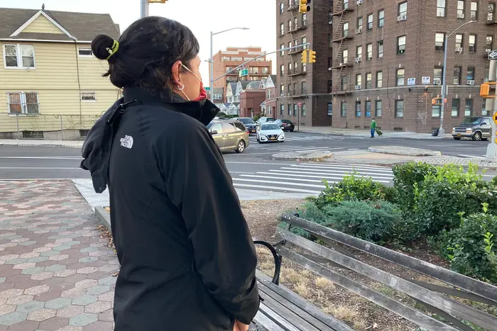 Pamela stands with her back to the camera, wearing a mask, facing a Queens street corner.